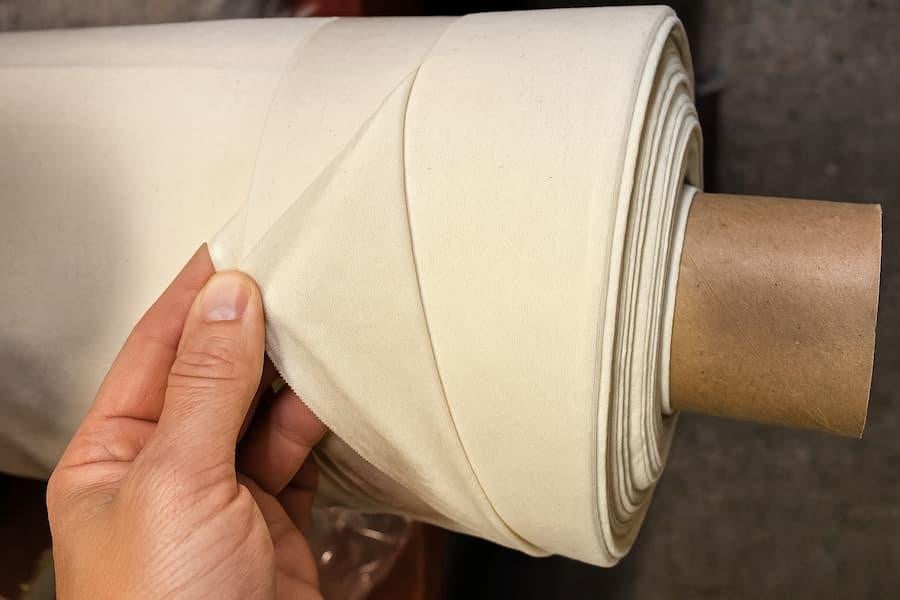 100% Cotton Muslin Lining Fabric (Organic), close-up of creamy white cotton muslin fabric on a roll being held by hand.