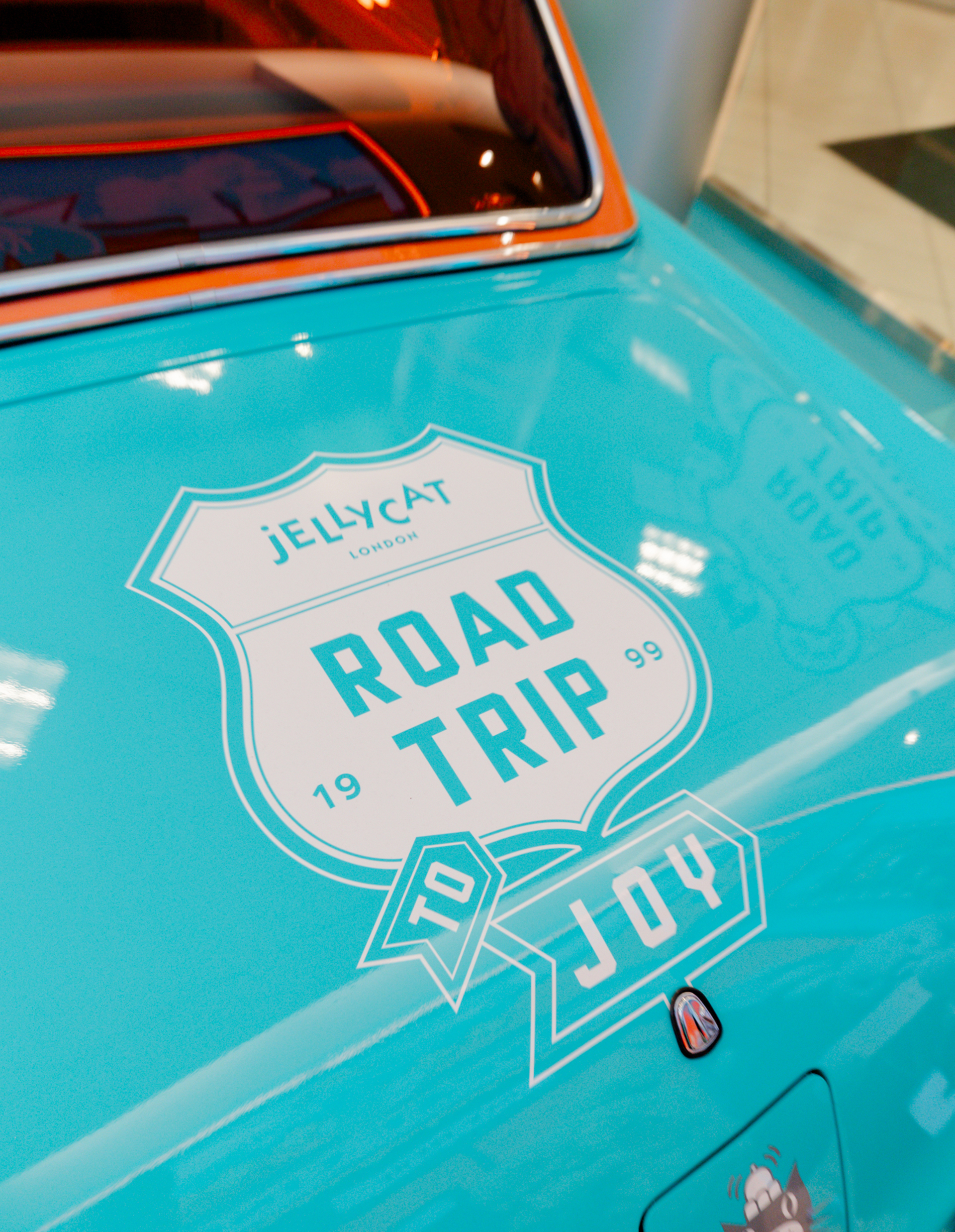 The hood of a vintage turquoise car features a Road Trip to Joy graphic inside a Jellycat-themed retail display.