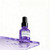 L'Oréal Professionnel Serie Expert Keratin Alpha Sleek Serum, 50 ml, in a purple spray bottle with a black cap, placed on a reflective purple surface with water droplets.