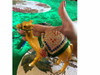 A handcrafted golden camel figurine adorned with a rich green saddle and vibrant red and blue jeweled embellishments.