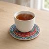 A professionally presented 9 cm round printed ceramic coaster featuring a unique Yin-Yang composition filled with intricate red and blue Persian arabesque rosettes and border illumination resting on a soft cream background.