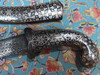 Product photo of the Handmade Engraved Metal Dagger 37 Cm configuration, showcasing an unsheathed ornamental weapon featuring a curved dark steel blade covered in tight, circular damascene-style textured patterns, paired with a matching silver-toned metal pistol-grip hilt and crossguard covered in dense, dark hand-engraved floral and leaf motifs, placed flat next to its coordinating silver metal sheath on a vibrant turquoise textile background woven with intricate pink, cream, and golden floral rug patterns.