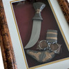 Product photo of the traditional Arabian Jambiya dagger inside its wall mount masterpiece frame, showcasing a wide, curved steel blade with an ornamented textured hilt mounted next to its matching silver and gold-toned scabbard on a deep burgundy velvet backing with a cream matting border, enclosed in a rectangular bronze-toned wooden frame isolated on a white background.