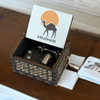 An open wooden hand-crank music box displaying mechanical inner workings and an illustration of a camel with an orange sun and "ABUDHABI" text on the inner lid, set against a wooden surface.