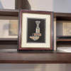Product photo of the traditional Arabian Khanjar dagger inside its 30x26 cm wall mount masterpiece frame, showcasing an intricately detailed silver-toned ceremonial dagger with ornate golden leaf scroll accents on the hilt and scabbard mouth, secured with a brown leather belt strap over a deep black matte background with a wide cream matting border, enclosed in a rectangular mahogany-finished wooden frame with an inner golden trim line, isolated on a white background.