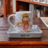 A professional product shot of the artistic winter ceramic mug and matching square saucer displayed on a polished dark wood table.