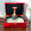 Product photo of the 16cm hand-painted copper vase, showcasing a slender teardrop silhouette with a flared polished copper neck and tiered pedestal base, highlighted by a central bulbous dark blue enamel band painted with a vibrant orange, red, and yellow central blossom medallion among green vines, displayed on top of a red velvet gift box with a gold clasp.