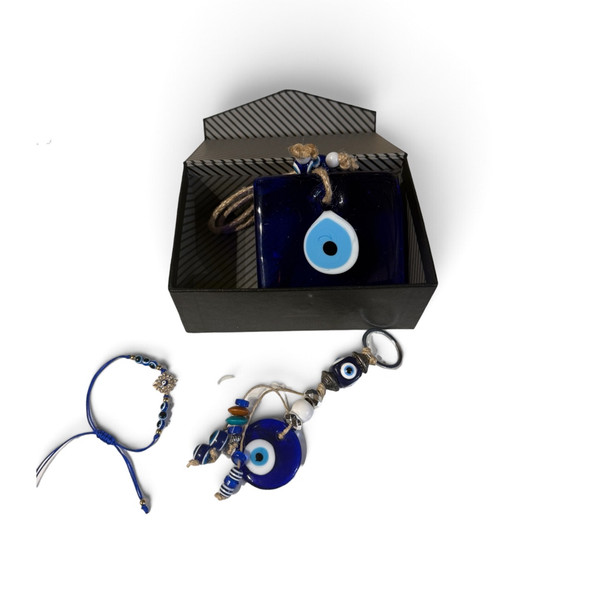 An overhead shot of an artisan Evil Eye protection gift set featuring a square glass evil eye wall hanging in a gift box, a blue cord bracelet, and a beaded keychain pendant with blue evil eye beads.