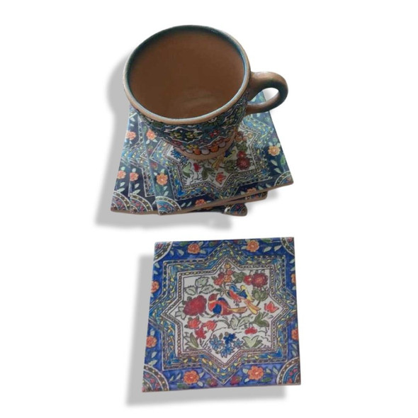 Close-up of a square ceramic coaster displaying intricate, colorful bird and floral artwork framed in an eight-pointed star against a blue patterned background.