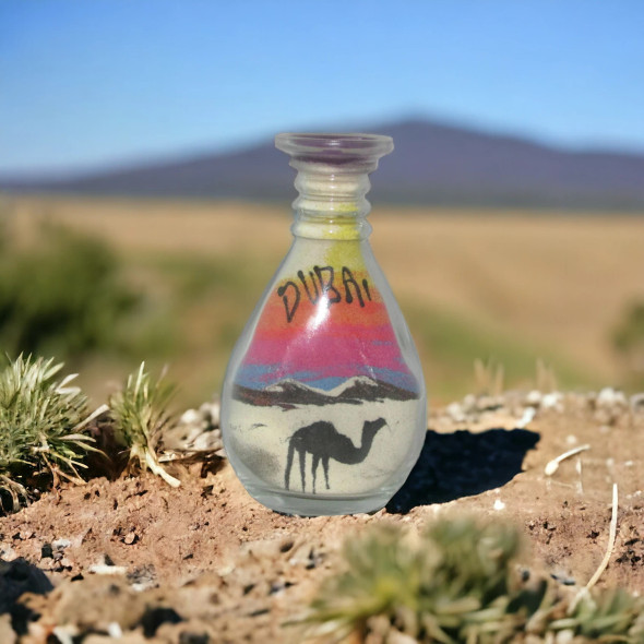 A flat small glass bottle featuring hand-poured sand art of a desert landscape with camels.