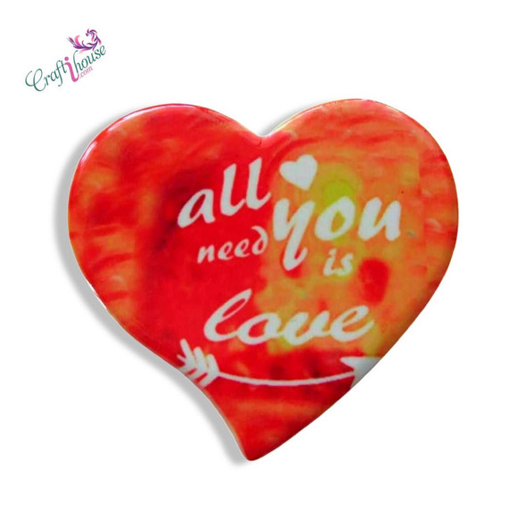 A heart-shaped printed ceramic fridge magnet featuring a vibrant red and orange watercolor background with the phrase "all you need is love" and a decorative arrow.