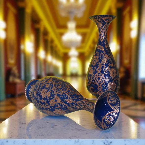 Product photo of the 25 cm copper engraved vase, showcasing a tall bulbous teardrop silhouette with a flared neck holding decorative dry sprigs, adorned with an intricate navy blue pattern of chiseled floral vine scrolls and geometric lattices, resting on a polished dark wood dining table with blurred red chairs in the background.