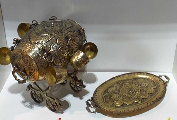 A professional product display shot of the ornate brass barrel dispenser with matching brass cups and engraved tray set on a woven chair.