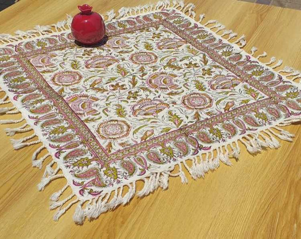 A 40x40 cm square hand-printed cotton table mat featuring traditional pink and olive-green paisley motifs, white fringes, and a decorative red pomegranate.
