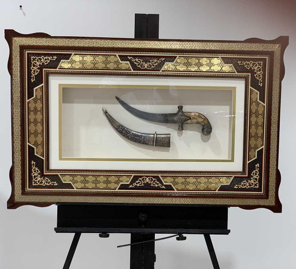 Macro view of the ceremonial weapon inside the shadow box, emphasizing the detailed chasing work on the crossguard, the natural grain patterns of the polished handle, and the textured finish on the curved decorative blade.