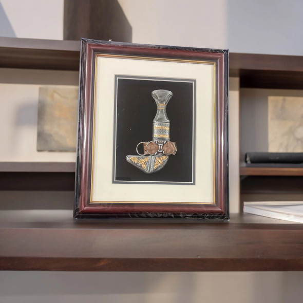 Product photo of the traditional Arabian Khanjar dagger inside its 30x26 cm wall mount masterpiece frame, showcasing an intricately detailed silver-toned ceremonial dagger with ornate golden leaf scroll accents on the hilt and scabbard mouth, secured with a brown leather belt strap over a deep black matte background with a wide cream matting border, enclosed in a rectangular mahogany-finished wooden frame with an inner golden trim line, isolated on a white background.