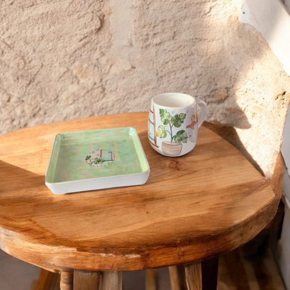A professional product shot of the artistic spring ceramic mug and matching square saucer displayed on a rustic wooden stool next to a potted plant.