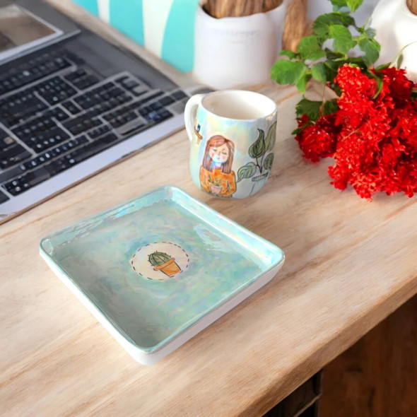 A professional product shot of the artistic plant lover ceramic mug and matching square saucer displayed on a wooden surface with bright red flowers.