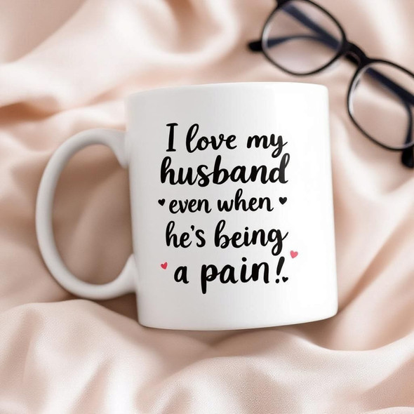 A clean, professional product photo of a "I love my husband..." mug set against a soft fabric background.