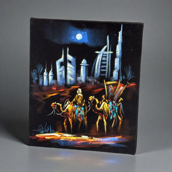 A 30x25 cm original oil painting on black velvet featuring twin camels glowing in amber light with the Dubai skyline behind.