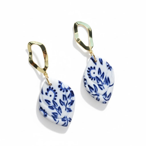 A pair of handcrafted leaf-shaped white clay earrings featuring a deep blue Chinoiserie floral pattern and hammered gold-toned open-link studs.