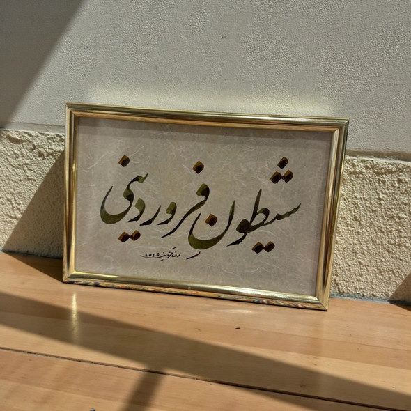 A 15.5 x 10.5 cm gold-framed Persian calligraphy piece featuring the phrase 'Farvardini Sheytoon' in black Nastaliq script on textured paper.