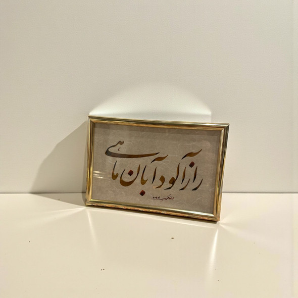 A framed Persian calligraphy piece with gold and dark brown script on textured paper, presented in a polished gold-toned frame.