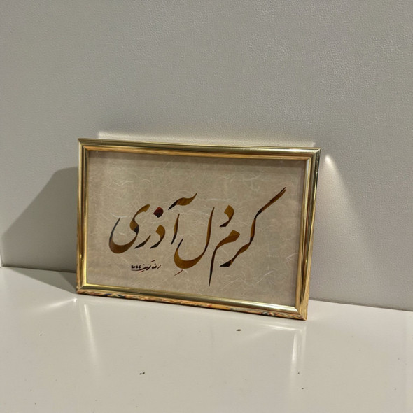 A framed Persian calligraphy piece featuring gold and umber script on textured paper, presented in a polished gold-toned frame.
