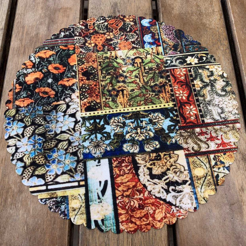 A 28 cm round velvet placemat on a rustic wooden table featuring a vibrant collage of multi-colored floral and botanical patterns.