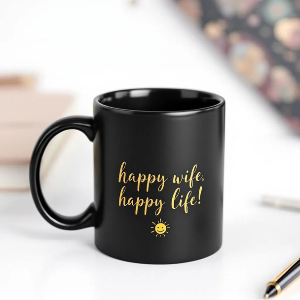 A professional product shot of a black ceramic mug with "Happy Wife, Happy Life" written in gold, set against a clean, modern workspace background.