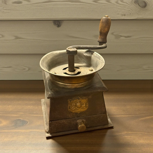 A side-angle view of a vintage manual coffee grinder featuring a weathered brass hopper, wooden handle, and antique metal body.