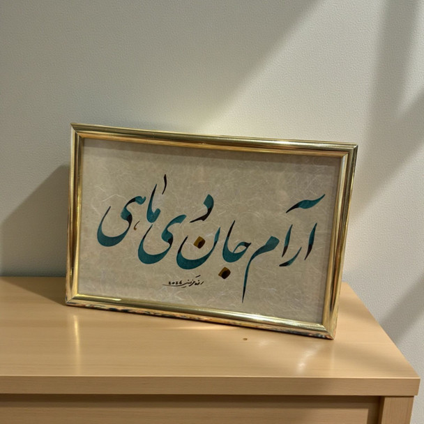 A framed Persian calligraphy piece with teal script and gold accents on a light-colored textured background, set in a gold frame.