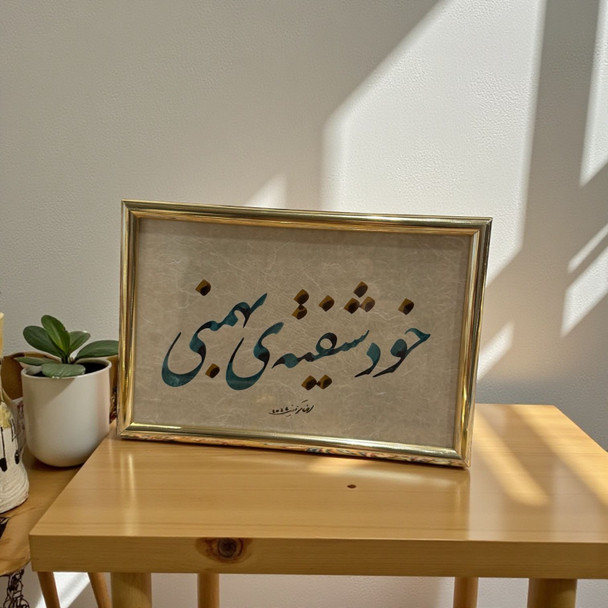 A framed Persian calligraphy piece featuring teal script and gold accents on textured paper, presented in a polished gold-toned frame.