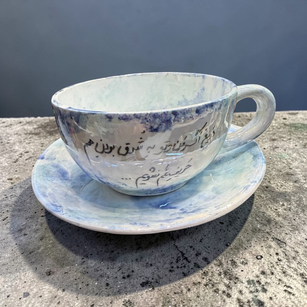 A handmade white ceramic cup and saucer set featuring sweeping black Persian calligraphy across the surface.