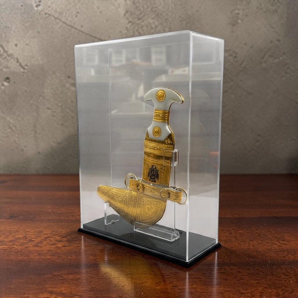 Product photo of the traditional golden color Khanjar dagger inside its acrylic display case, showcasing a beautifully sculpted ceremonial dagger with a white hilt and highly detailed golden sheath mounted vertically on a transparent internal stand, enclosed by a clear rectangular acrylic protective cover on a black base, styled on a polished dark wooden table against a textured concrete wall backdrop.