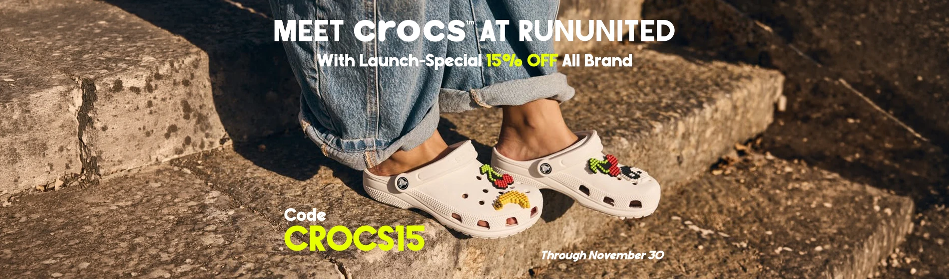 graphic, close-up of a person sitting on sunlit stone steps wearing blue jeans and white Crocs clogs decorated with colorful fruit Jibbitz charms, text, Meet CROCS at RunUnited with a Launch Special: 15% OFF All Brand Styles. Use code CROCS15 through November 30