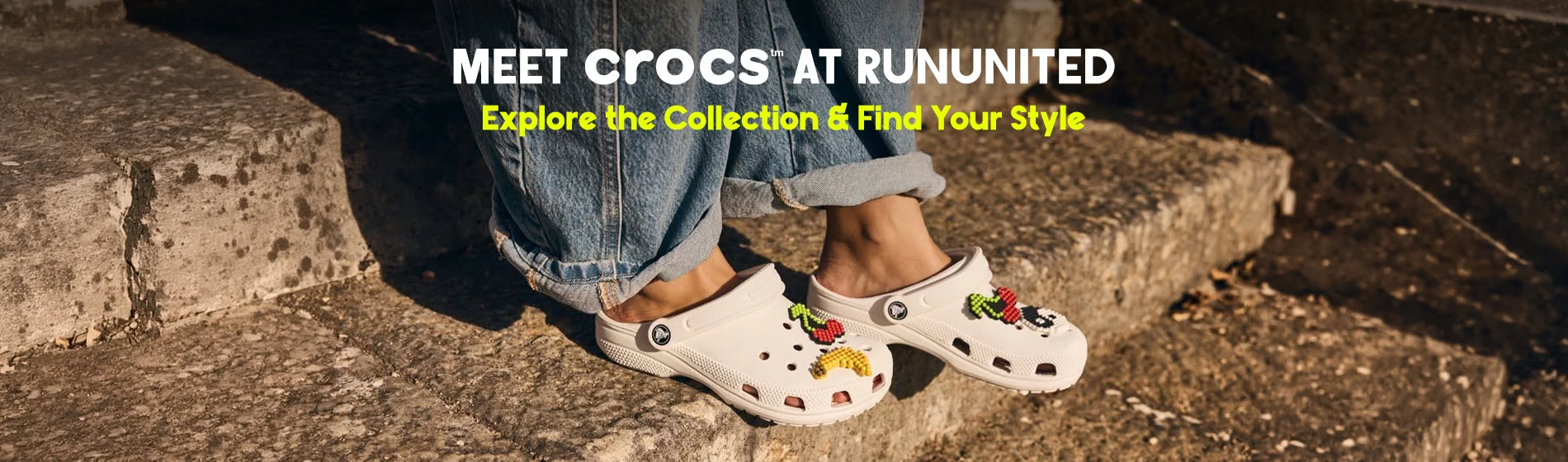 graphic, close-up of a person sitting on sunlit stone steps wearing blue jeans and white Crocs clogs decorated with colorful fruit Jibbitz charms, text, Meet CROCS Iconic Clogs: Explore the Collection & Find Your Style