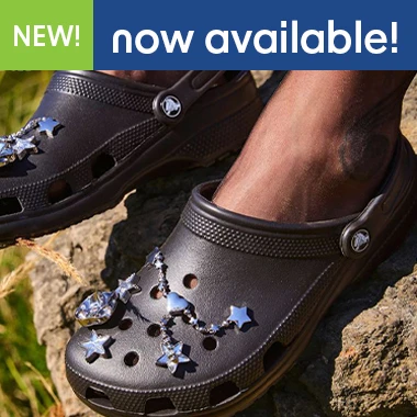graphic, close-up of a person's feet standing on a rugged rock wearing dark Crocs clogs decorated with star and dangling heart chain Jibbitz charms, text, Meet CROCS Iconic Clogs: Explore the Collection & Find Your Style