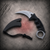 Silver fixed karambit knife with black sheath displayed separately on a textured gray background.