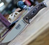 The ESEE-6 3D Handle (6PDT-005) shown with its 6.50-inch 1095 plain edge drop point blade in a Desert Tan finish and flat grind, 3D Coyote Brown and black G-10 handle scales with finger choil, and its injection-molded black plastic sheath.