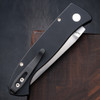 Close-up detail of the Pro-Tech Brend Classic BSA26.Brend.1 handle against a dark walnut wood background. The solid black aluminum scales, abalone button release, and satin hardware are the focal point of this shot.