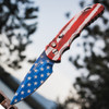 Pro-Tech TR-5 T501-PATRIOT shown deployed against an outdoor blue sky background. The USA flag Patriot Cerakote finish is clearly visible across both the handle and blade, with red and white stripes and blue field with white stars covering the full knife. The gold lip pearl push button and deep-carry pocket clip are visible on the handle.