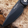 Pro-Tech TR-5 BSA26.TR5.1 shown open on a rough stone surface from a slightly different angle. The DLC black blade and feather-textured black aluminum handle are both visible, with the black lip pearl push button and black deep-carry clip in frame against the stone background.