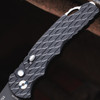 Close-up detail of the Pro-Tech TR-5 BSA26.TR5.1 handle against a dark background, showing the feather texture pattern milled into the black aluminum scales in full detail. The black lip pearl button release, slide safety, and satin hardware are clearly visible against the textured black aluminum surface.