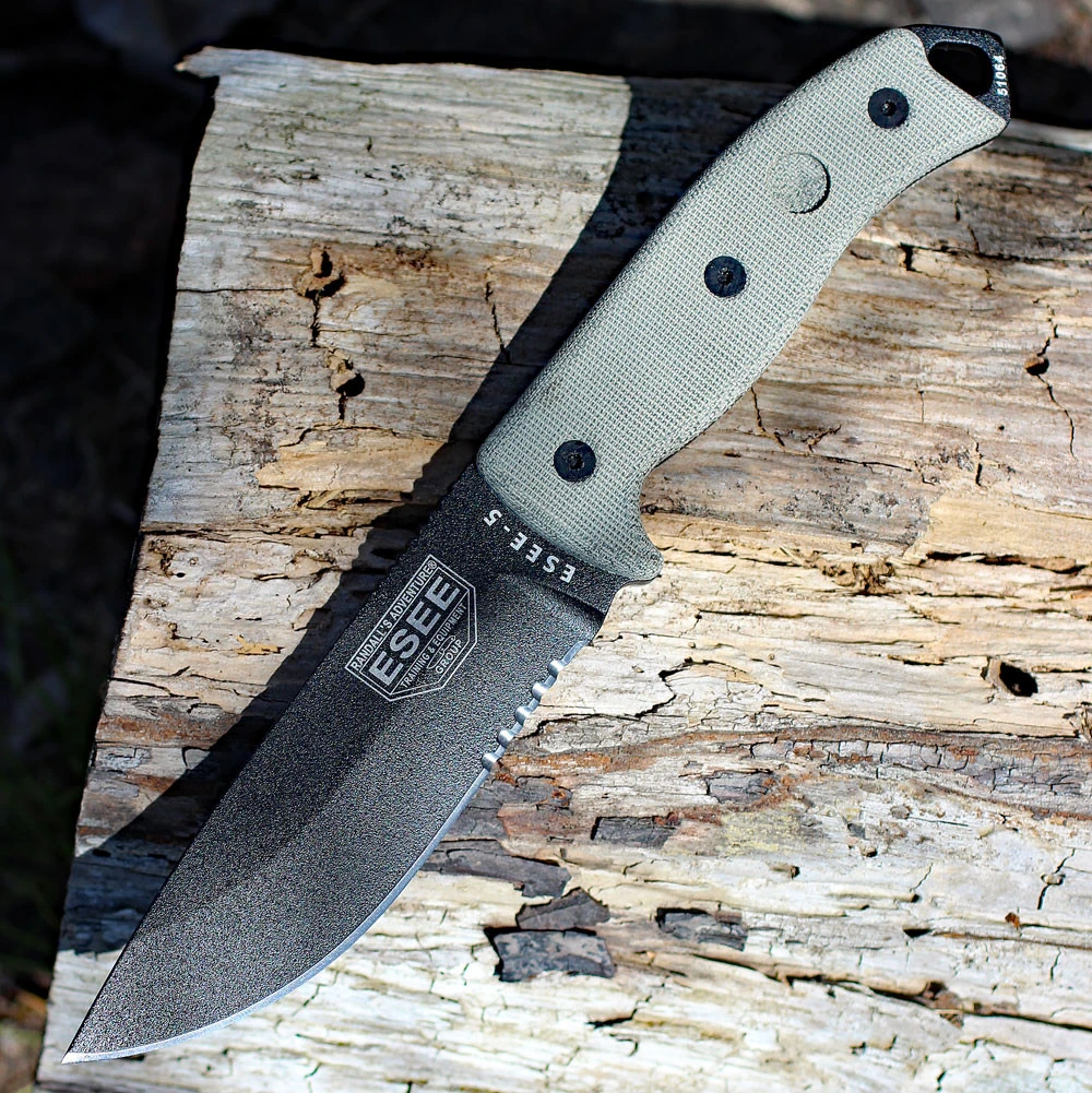 The ESEE-5 (ESEE-5S-E) shown with its 5.25-inch 1095 saber ground part serrated drop point blade in a black finish, sharpened glass breaker pommel, fire-making bow drill divot, tan Micarta handle scales, and its black Kydex belt sheath.