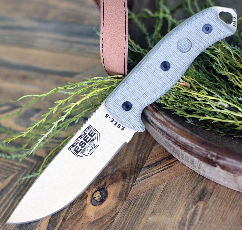 The ESEE-5 (ESEE-5P-DT) shown with its 5.25-inch 1095 saber ground drop point blade in a Desert Tan powder coat finish, sharpened glass breaker pommel, fire-making bow drill divot, tan canvas Micarta handle scales, and its black Kydex belt sheath.