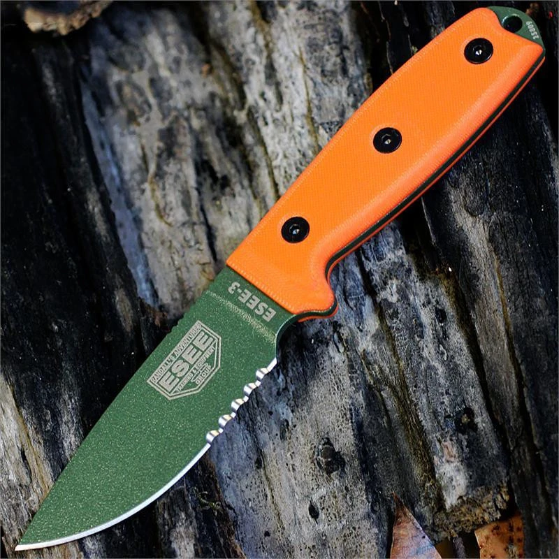 The ESEE-3 Fixed Blade (ESEE-3SM-MB-OD-CO) shown with its 3.88-inch 1095 carbon steel drop point blade in an OD Green finish with partial serration and flat grind, orange G-10 handle scales with a modified pommel, and its injection-molded black sheath with MOLLE back attachment.