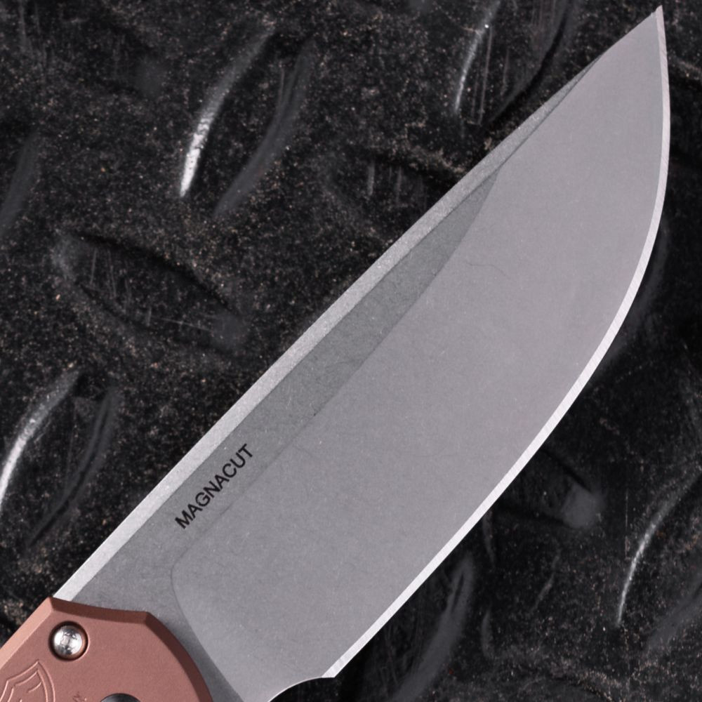 Pro-Tech Mordax BSA26.Mordax.1 shown open against a dark diamond plate background, angled to emphasize the stonewash CPM MagnaCut drop point blade. The blade's stonewash finish and flat grind are visible along the full length, with the brown aluminum handle and white pearl button visible at the base of the blade.