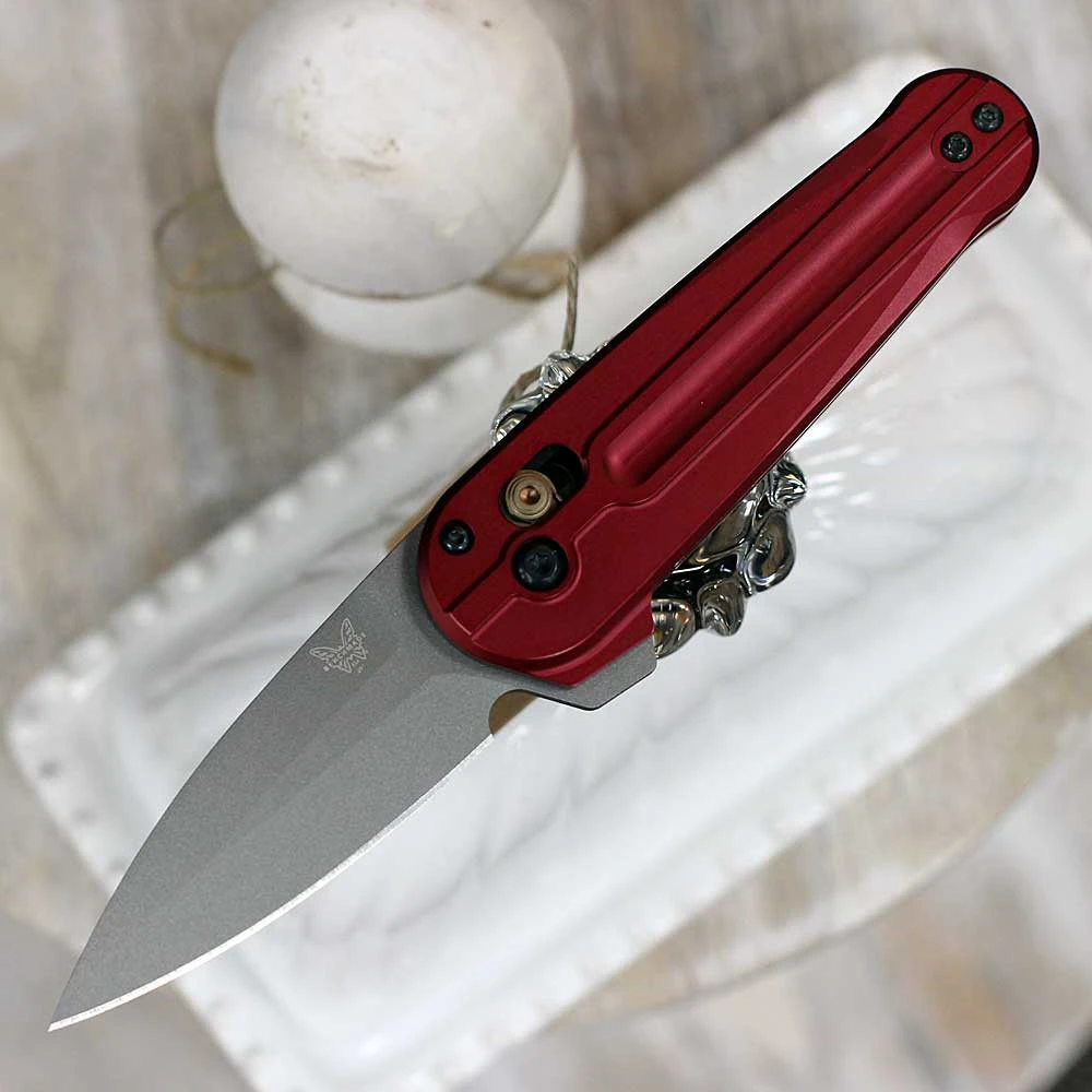 The Benchmade Lowden (491GY-01) shown with its 2.79-inch Böhler M390 spear point blade in a gray Cerakote finish and flat grind, milled Phoenix Red anodized aluminum handle scales, and its reversible tip-up pocket clip. The AXIS Lock flipper mechanism is visible along the spine of this compact, fidget-friendly EDC folder.
