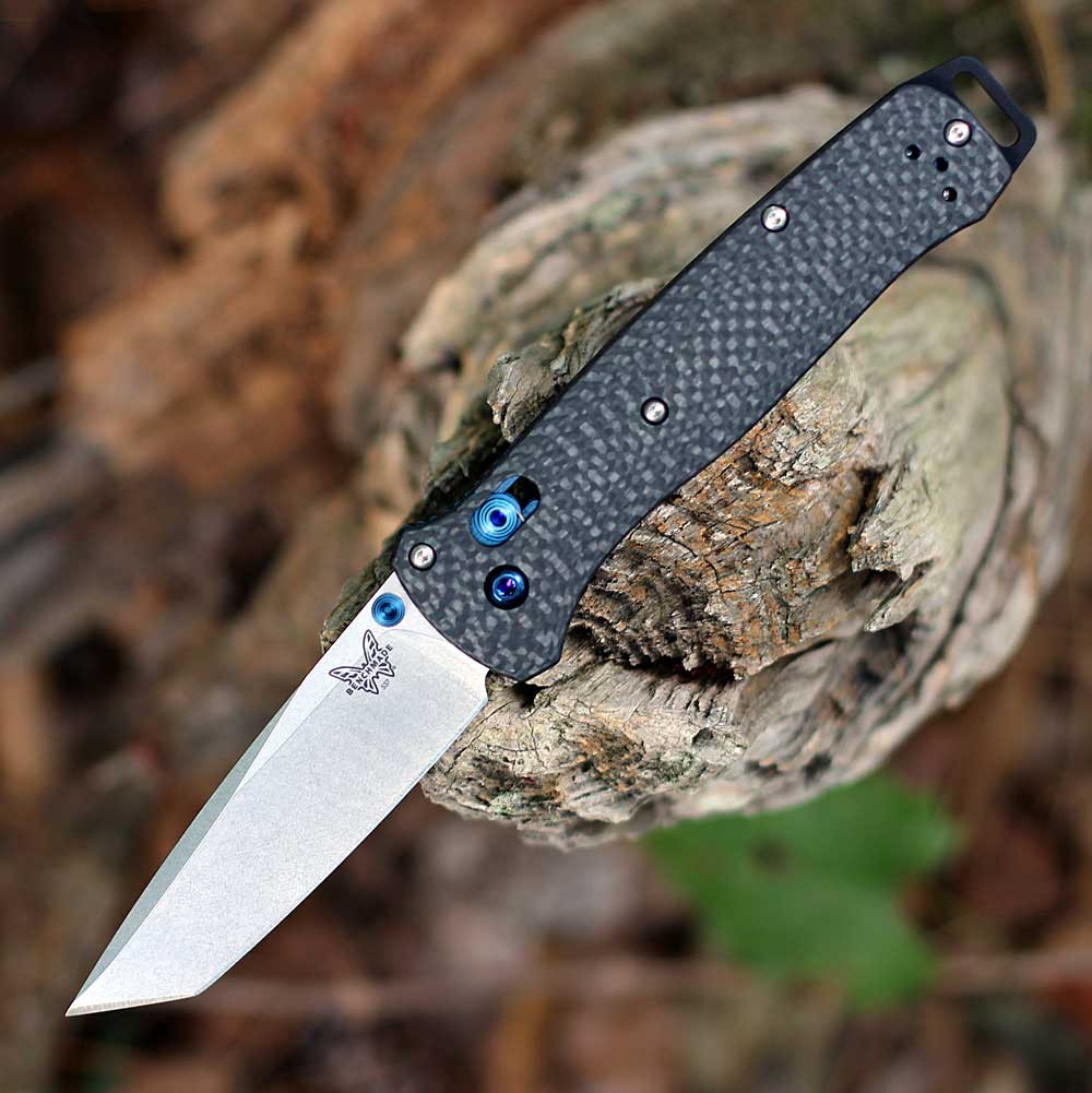 The Benchmade Bailout 537-09 is shown open, featuring a 3.40-inch Stonewashed Böhler M390 tanto blade extending from a contoured Carbon Fiber handle with Blue anodized pommel and hardware accents, dual thumb studs, and AXIS Lock mechanism.